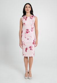 Ärmelloses Kleid aus hellrosa Stoff mit einem floralen Muster in Rot und Weiß, mit einer taillierten Silhouette und knielangem Saum. Silberne Schuhe.
