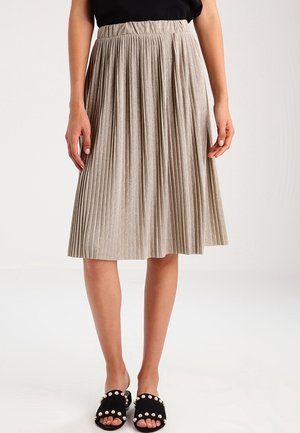 Femme portant une jupe plissée beige longueur genoux, un haut noir sans manches et des sandales noires ouvertes aux orteils avec des décorations en perles.