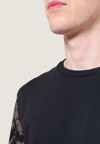 Czarna bluza z okrągłym dekoltem, zdobiona teksturowanymi brązowymi abstrakcyjnymi wzorami na rękawach. Miękki materiał bawełniany, dopasowany krój.