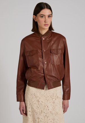 Junge Frau trägt eine braune Lederjacke mit Fronttaschen und einen beigen, floralen Spitzenrock vor einem einfarbigen Hintergrund.