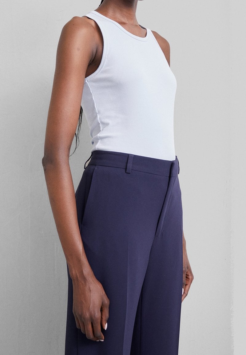 Personne portant un haut sans manches blanc côtelé et un pantalon sur mesure taille haute bleu foncé sur un fond uni.