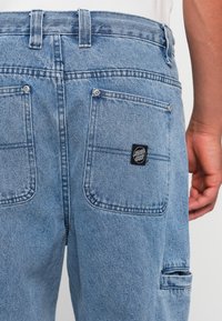 Lichtblauwe spijkerbroek met een hoge taille, dubbele achterzakken en een zwart label logo op de rechterzak. Zachte textuur met stiksels.