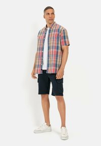 Korte mouwen geruite shirt in rood, blauw en beige, gecombineerd met donkere cargoshorts. Model draagt witte sneakers en een effen shirt eronder.