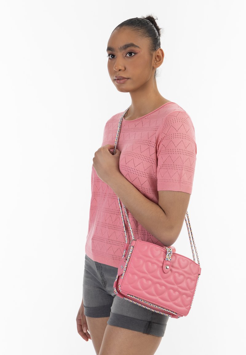 Rosa vadderad handväska med hjärtformade mönster, kedjestropp och en texturerad yta, bärs med en matchande rosa stickad topp och grå shorts.