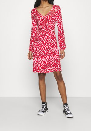 Woman wearing a red, long-sleeve, knee-length dress with white daisy patterns and black high-top sneakers.