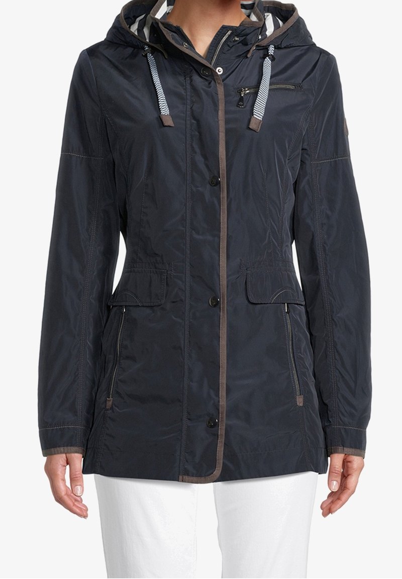 Veste à capuche bleu marine foncé avec bordure marron, fermeture éclair et boutons à l'avant, poche poitrine zippée, rabat et poches latérales zippées, portée sur un pantalon blanc.