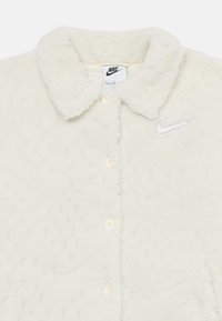 Krēmkrāsas šallīte ar teksturētu virsmu, ar apkakli, pogām un sudraba Nike logo uz krūtīm.
