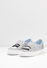 Silver glittriga slip-on-sneakers med broderade ögon, ett öppet och ett som blinkar, på vita gummisålar mot en vit bakgrund.
