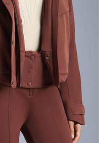 Barna, testhezálló sportdzseki cipzárral és patentgombokkal, krémszínű bordázott felsővel rétegezve, hozzáillő barna leggingsszel párosítva.