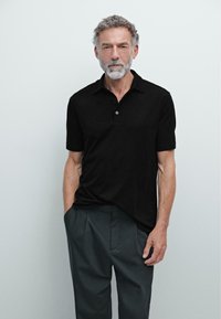 Mand med gråt hår og skæg iført en sort kortærmet polo-shirt og mørkegrå bukser stående mod en ensfarvet lys baggrund.