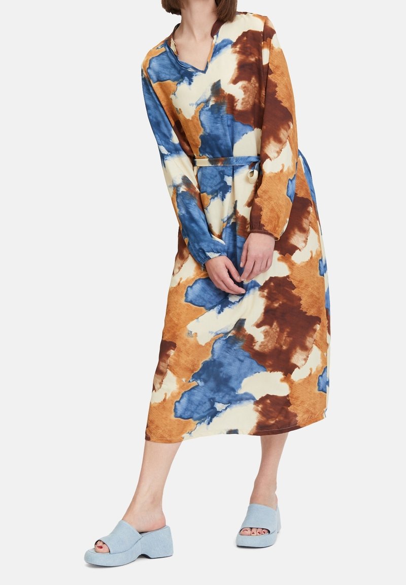 Femme portant une robe à manches longues ceinturée avec des motifs abstraits bleus, bruns et crème, et des sandales à plateforme bleu clair à bout ouvert.