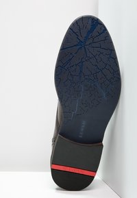 Sole di scarpa in pelle marrone con fondo in gomma navale testurizzato caratterizzato da un motivo screpolato. Accento rosso sul tallone. Dettagli laterali lisci.