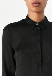 Svart langermet skjorte med subtil tekstur og tonalt mønster. Har krage, knappefeste, og en dekorativ knapp ved halsen.