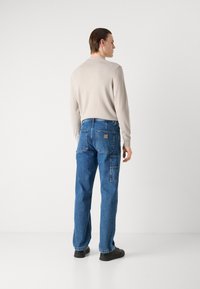 Camisa de manga larga beige combinada con vaqueros de denim azul con bolsillos y parche de marca, llevada con zapatillas negras.