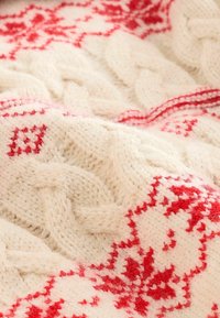 Gros plan sur un tissu en tricot crème avec des motifs de flocons de neige et de losanges rouges, avec de grosses torsades et une texture douce et duveteuse.