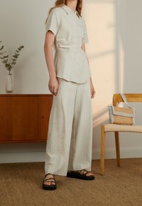 Il modello indossa una camicia a righe leggera e pantaloni larghi, in piedi accanto a un mobile in legno e a una sedia con una borsa intrecciata sopra.