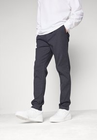 Tmavě šedé kalhoty na míru z texturovaného materiálu s přímým střihem a bočními kapsami, kombinované s bílými teniskami.