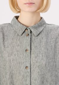Camicia di lino grigio chiaro con colletto a punta, chiusura a bottoni con bottoni marroni, tessuto testurizzato con variazioni sottili.