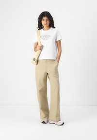 T-shirt blanc à manches courtes avec un graphique "Carhartt WIP", pantalon large kaki, et sac bandoulière beige. Le modèle porte des baskets roses.