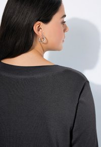 Femme aux longs cheveux noirs portant un haut en tricot gris foncé et des boucles d'oreilles en argent, de profil contre un fond clair.
