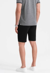 Schwarze Baumwollshorts mit seitlichen Taschen, kombiniert mit einem grauen Polohemd mit einem schwarz gestreiften Kragen. Getragen mit weißen Slippers.