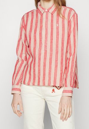 Femme portant une chemise à manches longues rayée verticalement rouge et beige, boutonnée, associée à un pantalon blanc avec un petit motif brodé.