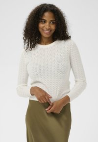 Femme souriante aux cheveux bouclés, portant un pull long à manches longues texturé blanc et une jupe olive, se tenant devant un fond clair uniforme.