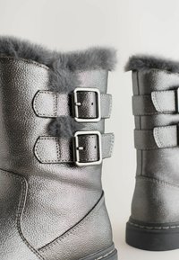 Sudraba teksturēti ziemas zābaki ar pelēku kažokādu un dubultiem jostu siksniņām, redzami no aizmugures uz baltas fona.