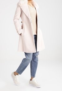 Manteau rose clair avec un col en fausse fourrure, superposé à un pull beige, associé à un jean bleu déchiré et des baskets beiges.