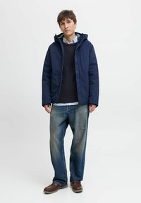 Navy padded jacket with hood, gray knit sweater, and light blue striped shirt beneath; paired with loose-fitting denim jeans and brown shoes.