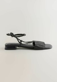Mustad mustad sandaalid, millel on kootud tekstiilist pealis, sirge tald ja punutud pahkluu rihm madala plokk-kandiga.