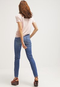 Frau mit roten Haaren, die ein hellrosa T-Shirt, enge blaue Jeans und braune Plateausandalen trägt, steht mit einer Hand auf ihrem Rücken.