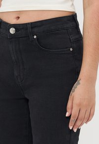 Černé džíny z denimu s přiléhavým designem, s vysokým pasem, stříbrným knoflíkovým zapínáním a předními kapsami. Hladká textura a klasický styl.