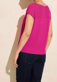 Fuksiansävyinen yläosa, jossa lyhyet neulotut hihat ja teksturoitu kuviointi. Takana sileä pinta ja väljä istuvuus, yhdistetty tummansinisiin farkkuihin.