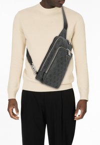 Gray sling bag with a textured finish, multiple zippered compartments, and a black adjustable strap, worn across the body over a beige sweater.