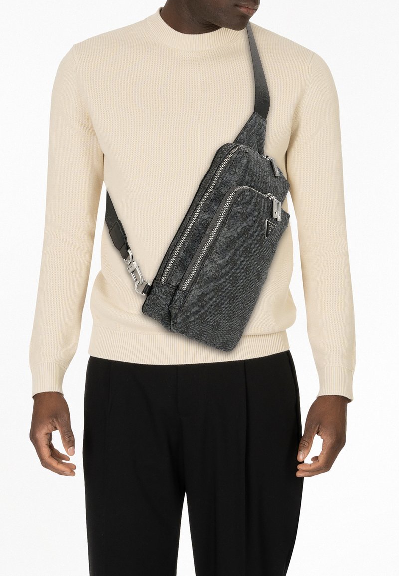 Gray sling bag with a textured finish, multiple zippered compartments, and a black adjustable strap, worn across the body over a beige sweater.