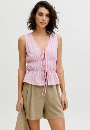 Femme aux cheveux bouclés et foncés portée un haut rose rayé sans manches avec des liens et un short beige ample, debout devant un fond clair uni.