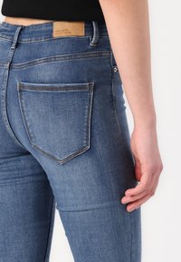 Blugi subțiri din denim albastru, cu talie medie, design cu cinci buzunare și cusături distincte la buzunarul din spate, având un eticheta de marcă maro.