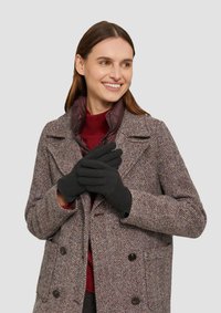 Schwarze Handschuhe, die von einer Person in einem mantel mit Fischgrätenmuster in Braun-, Rot- und Creme-Tönen und einem kastanienbraunen Pullover darunter getragen werden.