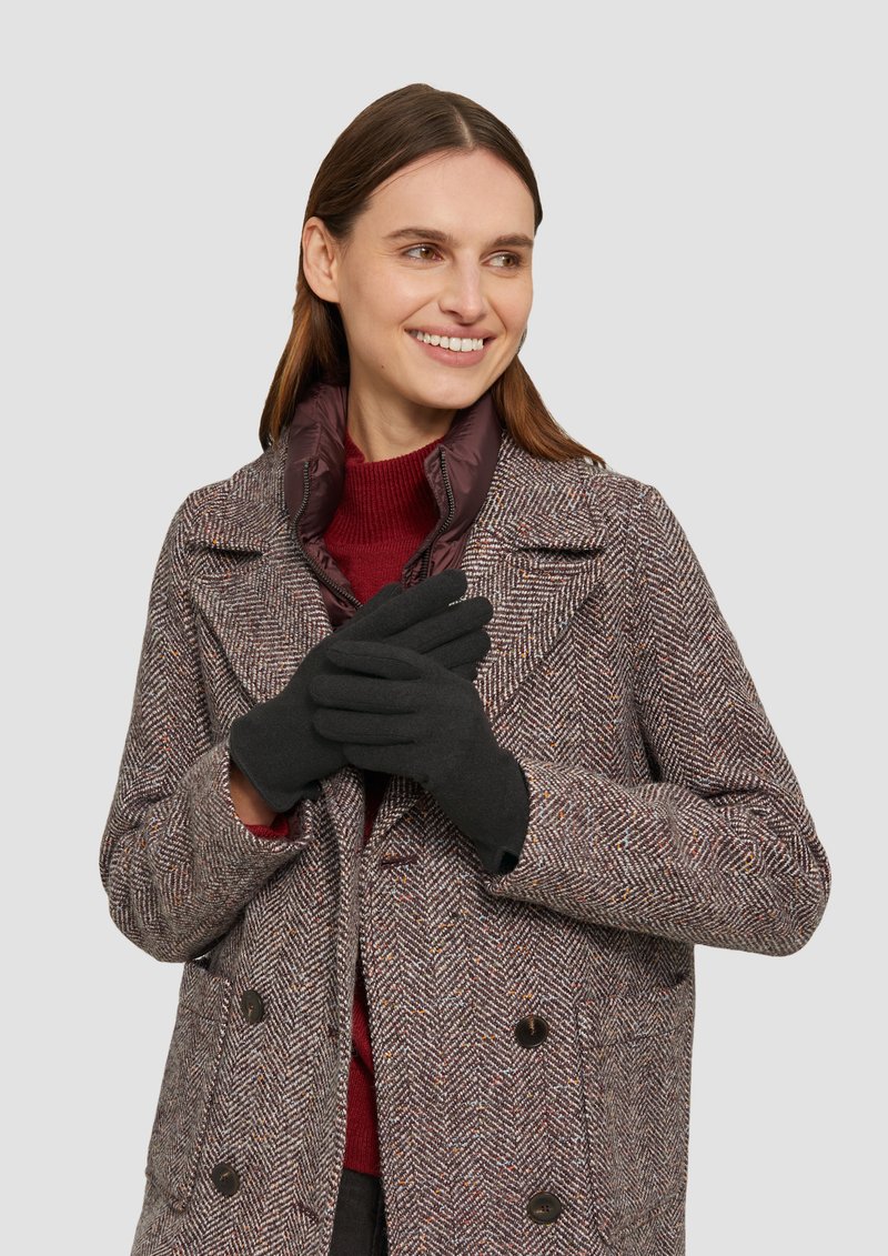 Schwarze Handschuhe, die von einer Person in einem mantel mit Fischgrätenmuster in Braun-, Rot- und Creme-Tönen und einem kastanienbraunen Pullover darunter getragen werden.