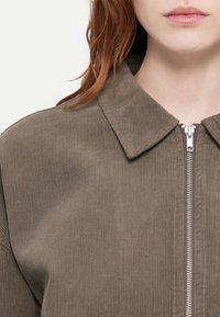 Bruine corduroy jas met een kraag en een zilveren ritssluiting aan de voorkant. De stof heeft een verticale geribbelde textuur, wat zorgt voor een zachte uitstraling.