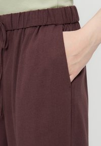 Χέρι που γλιστρά στην πλαϊνή τσέπη ενός φαρδιού, σκούρου καφέ παντελονιού με ελαστική μέση και κορδόνι, φορώντας ένα ανοιχτό πράσινο τοπ.