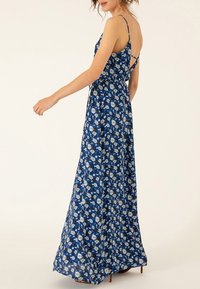 Robe maxi avec des bretelles fines, en tissu bleu orné de motifs floraux blancs, silhouette en A, et un dos ouvert plongeant.