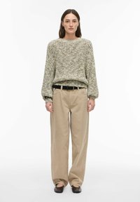 Pull en maille épaisse texturée de couleur beige clair avec larges poignets ; associé à un pantalon kaki ample taille haute et une ceinture noire.