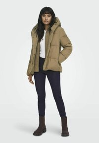 Béžová zateplená bunda s kapucí, s patentkami a prošívaným designem, nošená s bílou košilí a tmavými džínami, doplněná hnědými kotníkovými botami.