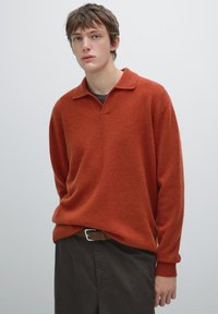 Sweter w kolorze rdzy wykonany z miękkiej przędzy, z kołnierzykiem, ściągaczami przy mankietach i luźnym krojem. W zestawieniu z ciemnymi spodniami.
