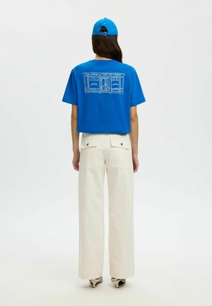 Femme portant une casquette bleue, un t-shirt bleu avec un croquis de devanture blanche, et un pantalon ample écru, debout de dos sur un fond blanc.