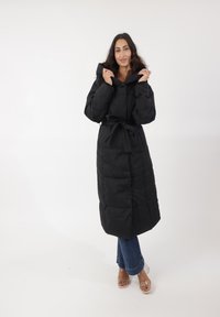 Abrigo acolchado negro largo con cinturón, que presenta una capucha, diseño acolchado y tejido texturizado. La modelo lleva jeans de mezclilla y sandalias transparentes.