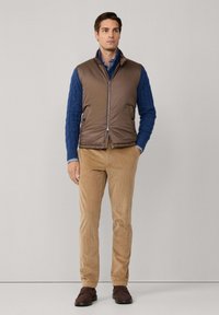 Gilet imbottito marrone sopra un maglione blu a trecce, abbinato a pantaloni in corduroy beige e scarpe mocassino marroni. Design semplice e funzionale.