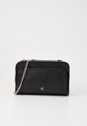 Sac bandoulière en cuir noir avec anse chaîne argentée et poche avant, arborant un petit logo "cK" sur un fond blanc uni.